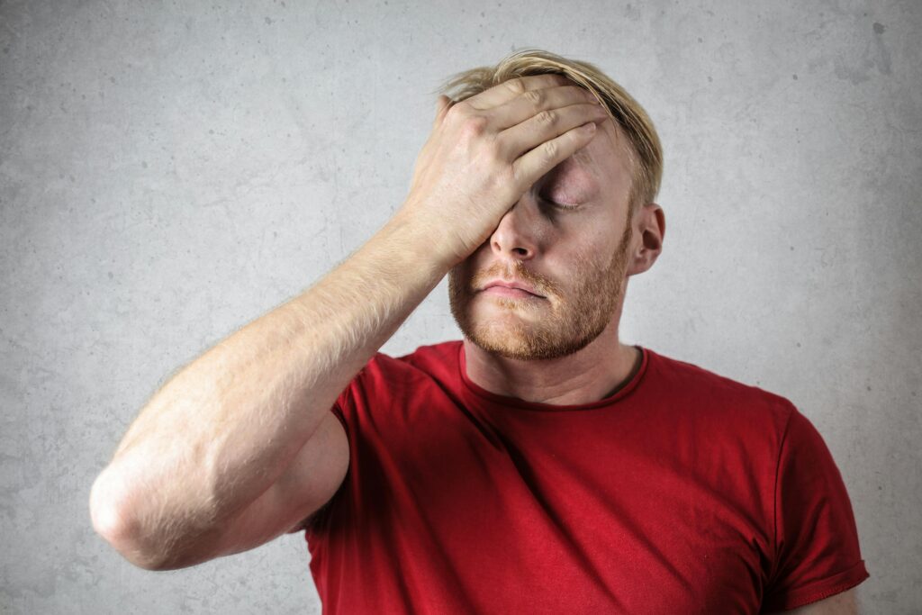 Man in red shirt looking confused and frustrated, covering his face with his hand, representing the difficulty in choosing between alkaline water and purified water for optimal health benefits.